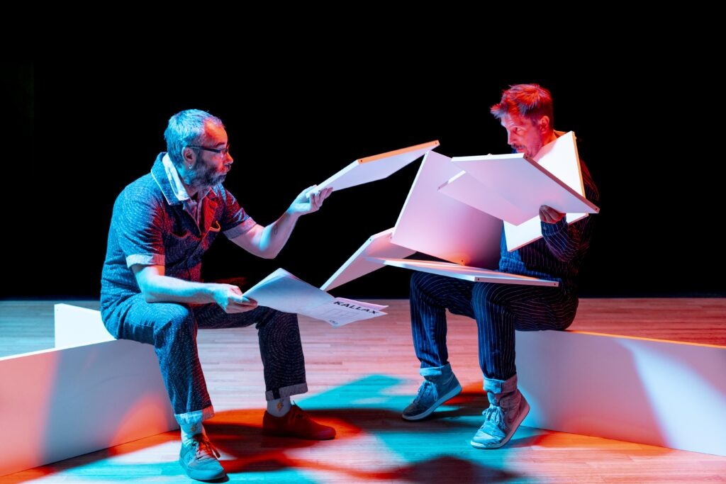 Deux hommes sur scènes assis avec une lumière bleu et rouge, essayant de comprendre une notice pour monter un meuble. Spectacle Pling-Klang